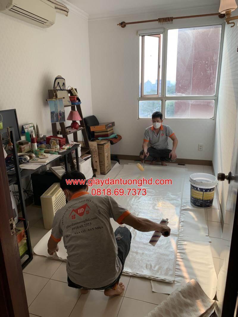 Hai thợ thi công đang trải giấy dán tường ra sàn để lăn keo chuyên dụng trước khi dán lên tường phòng khách căn hộ.