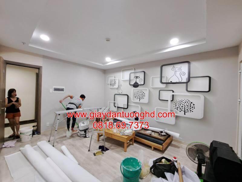 Thợ thi công đang lắp đặt giấy dán tường trơn màu xám nhạt hiện đại làm nền cho vách trang trí kệ gỗ phòng khách.