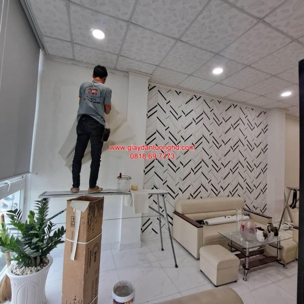 Thợ đang đứng trên giàn giáo tỉ mỉ cân chỉnh và thi công giấy dán tường màu trắng trơn bên cạnh mảng tường họa tiết hình học ziczac đen trắng.